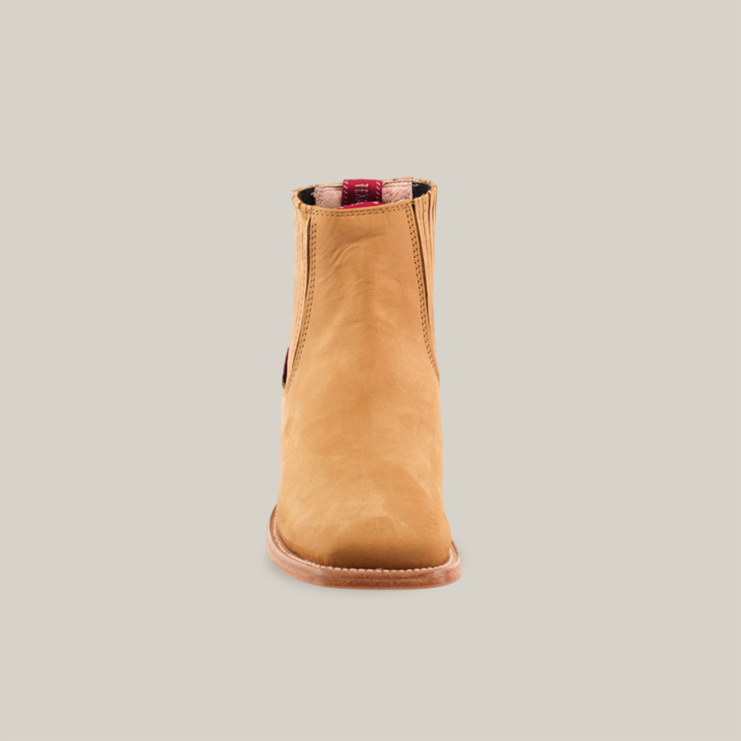 The Prime Suede Honey Square Toe, a single tan leather ankle boot with a hint of cowboy flair, features a brown sole. Seen front-on against a white background, it boasts a stylish red and black pull tab at the top of the opening.