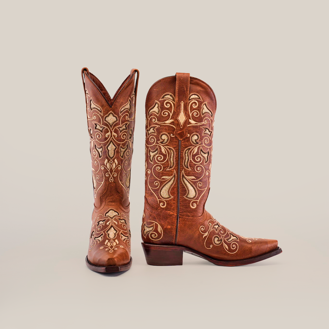 The Nataly Brown Mid Shaft Snip Toe boots, with expert artisan craftsmanship, boast premium leather and detailed white and gold embroidery. Displayed on a plain white background, one boot stands upright while the other lies on its side, showcasing their exquisite patterns.