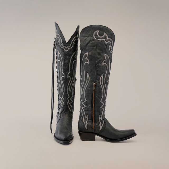 A pair of handcrafted Tania Matte Black cowboy boots, tall with a snip toe design and adorned with intricate white stitching. Featuring side zippers and decorative embroidery, the left boot stands upright while the right leans slightly to the side.