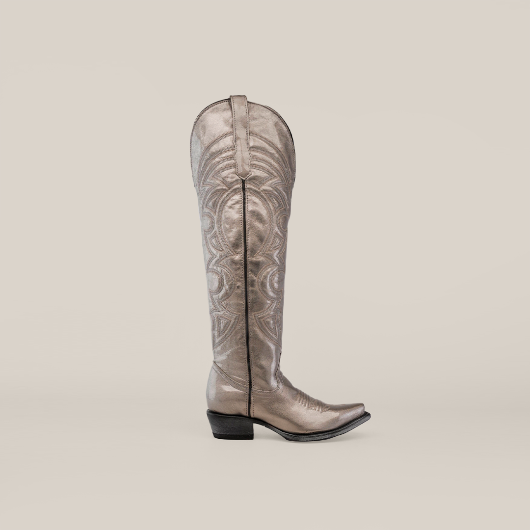 Against a plain gray background, the Taylor Mercury - Tall Shaft - Snip Toe cowboy boot stands out with intricate swirling patterns, a metallic silver finish, and a leather sole, showcasing elegance and craftsmanship.