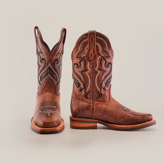 The Fresno Shedron boots feature a stylish rodeo toe with intricate black and brown stitching on the shafts. One boot stands upright while the other is angled to reveal its side pattern and slip-resistant sole, all set against a white background.