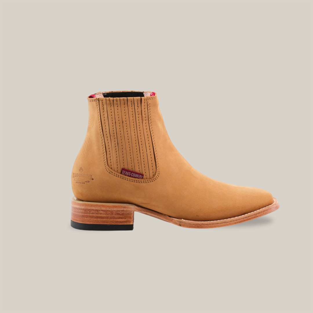 The Prime Suede Honey Square Toe boot embodies cowboy flair with its tan leather, sleek design, red inner lining accents, vertical side stitching, tag by the elastic gussets, wooden low heel, and black sole.