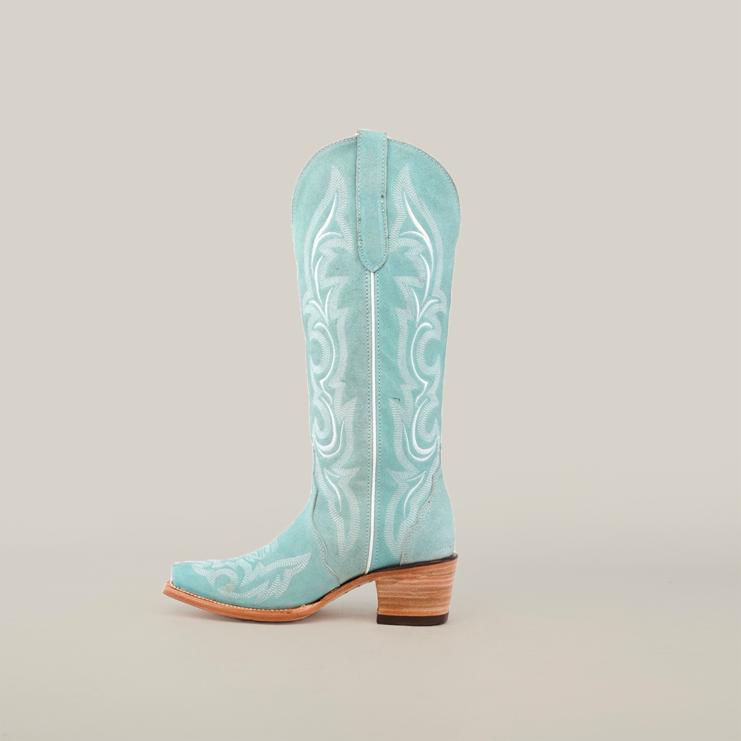 The Amara BB Suede features a single light turquoise western boot with an elegant snip toe, intricate white stitching on its tall shaft, and a stacked wooden heel, set against a plain white background.