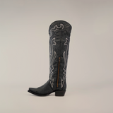 The Tania Matte Black boot, featuring a tall shaft and snip toe, is showcased against a plain background. This handcrafted boot boasts a matte black finish with ornate white stitching, a side zipper, low heel, and intricate western-style designs.