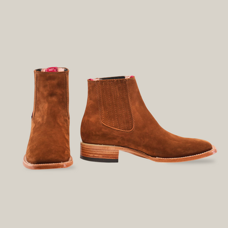 The Prime Suede Caramel Square Toe boots, with a Western touch and wooden soles, are featured on a white background. One boot stands upright and the other is turned to display intricate stitching, emphasizing this cowboy fashion staple.