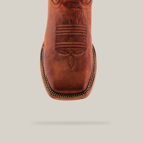 The Rustic Cogñac - Square Toe is a handcrafted boot made of cognac cowhide leather, featuring intricate stitching, a square toe, and decorative patterns. It stands upright against a white background, highlighting its craftsmanship and elegance.