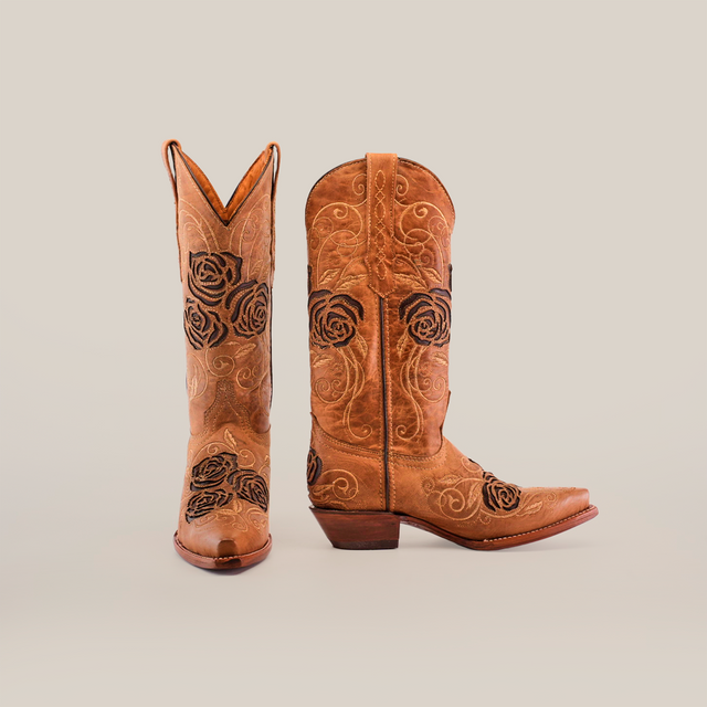 These Rosal Fawn Mid Shaft Snip Toe boots embody handcrafted elegance with premium leather, one showcasing its brown hue and black embroidered roses upright, while the other is angled to reveal side detailing against a white backdrop.