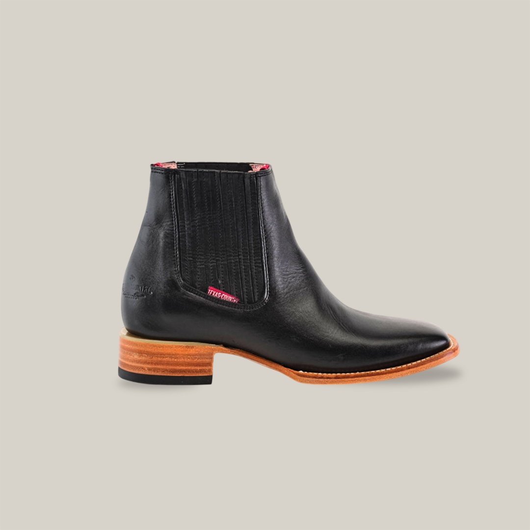 The Napa Black Square Toe is a handcrafted black leather ankle boot with a wooden sole and small heel. It features elastic side panels for easy slip-on, a subtle red tag, and its sleek light brown finish adds a hint of cowboy style.