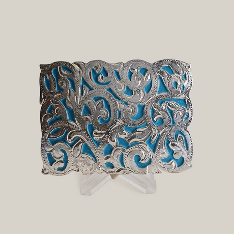 The Charro Turquoise Buckle is an ornate alpaca silver piece featuring intricate scrolling patterns on a vibrant blue background. Its cut-out design showcases exceptional craftsmanship with highlighted curves and flourishes, reminiscent of classic Texas-style buckles, displayed upright on a stand.