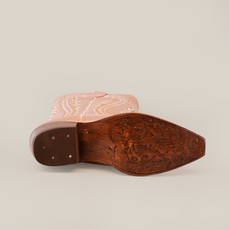 The image displays the bottom view of a tan cowboy boot on its side, showcasing a snip toe with an ornate carved sole featuring floral and abstract patterns. Light-colored decorative stitching along the side mirrors the design of Sara Metalic Florentine - Tall Shaft - Snip Toe boots.