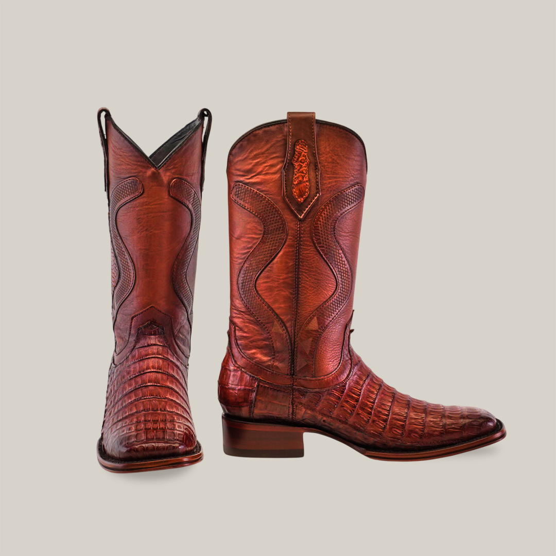 A pair of Exotic Caiman Belly Tail - Diamond Brandy - Square Toe cowboy boots, crafted from caiman belly leather, beautifully showcase intricate stitching and a pointed toe. Displayed gracefully on a smooth white background, one stands upright while the other is slightly tilted.