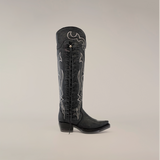 The Tania Matte Black boot offers a knee-high western style with decorative embroidery, side lace-up detail, snip toe, and low heel. Handcrafted craftsmanship shines against a plain, light grey background.