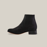 The Prime Suede Black Round Toe is a sleek black suede ankle boot with a low wooden heel. It features an elastic side panel for easy wearing and a round toe design, perfect for a cowboy-inspired ensemble, all showcased on a plain white background highlighting its smooth finish.