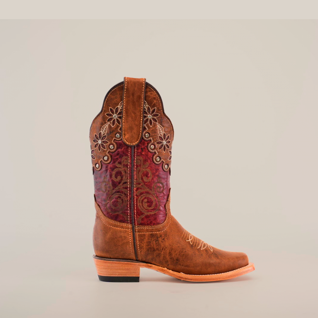 The Grecas Orix boot features a brown leather exterior and decorative upper with red floral embroidery and intricate patterns. It has a short shaft, narrow square toe, and low wooden heel, all set against a plain white background.