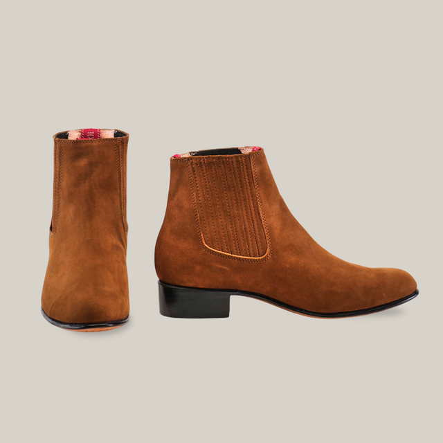 The Prime Suede Caramel Round Toe boots are handcrafted with brown suede, feature slight heels, elastic side panels, and a red plaid lining. One boot shows the back while the other displays a stylish side profile, exuding classic Western charm.