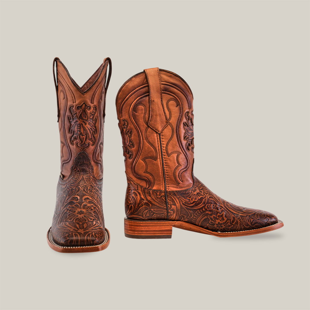 The Handtooled Leave Brown cowboy boots, crafted from premium cowhide leather and featuring ornate stitching, are showcased with the left boot upright and the right one tilted to reveal square toe detailing against a neutral background.