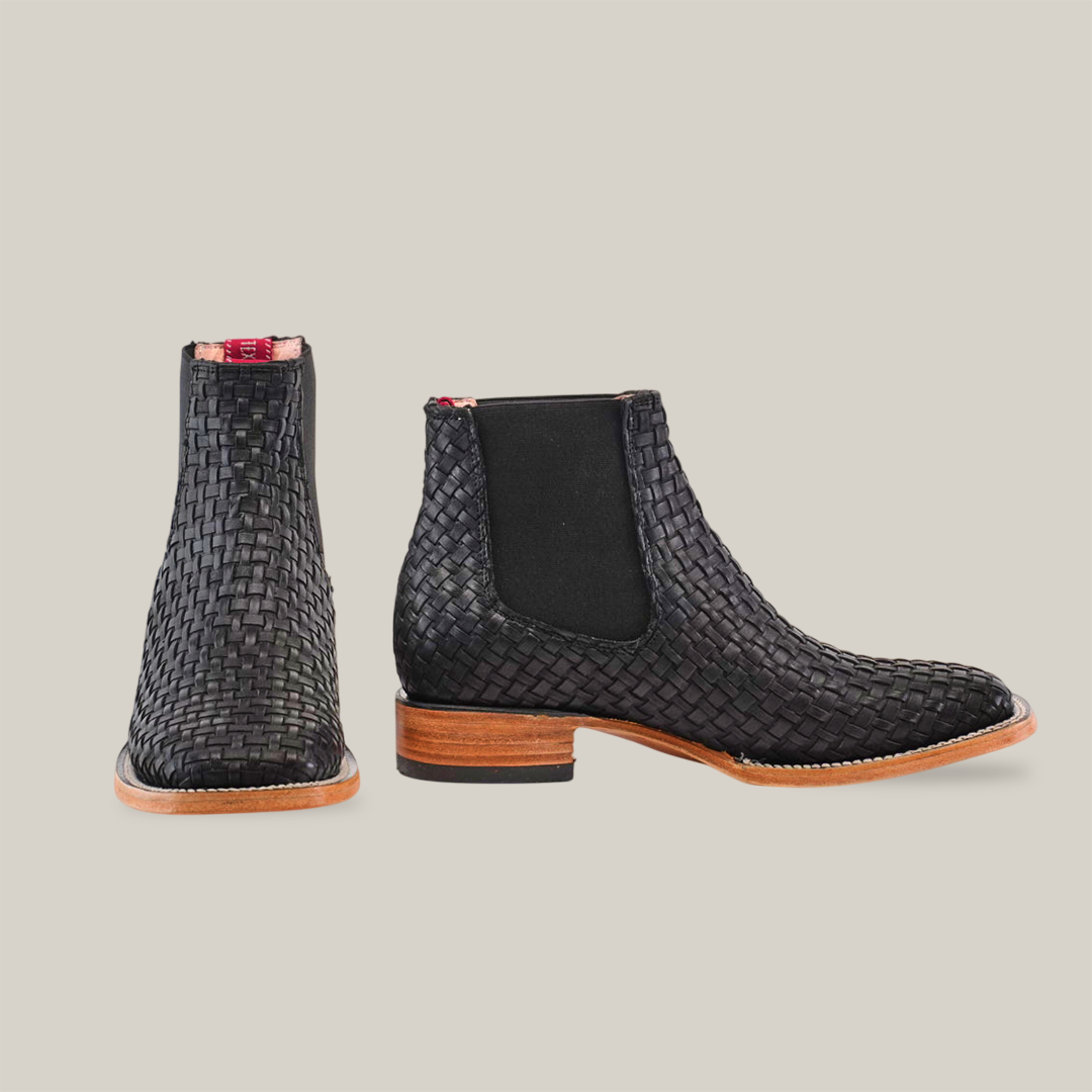 The Basket Weave Black Square Toe ankle boot features a chic basket weave design, square toe, brown wooden heel and sole, black elastic side panels, and a playful pink back tab. Set on a crisp white background, it exudes style and sophistication.